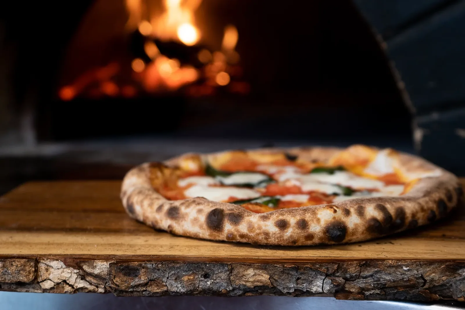 A Neapolitan-style Margherita pizza with blistered crust on a wooden board, in front of a glowing wood-fired oven.